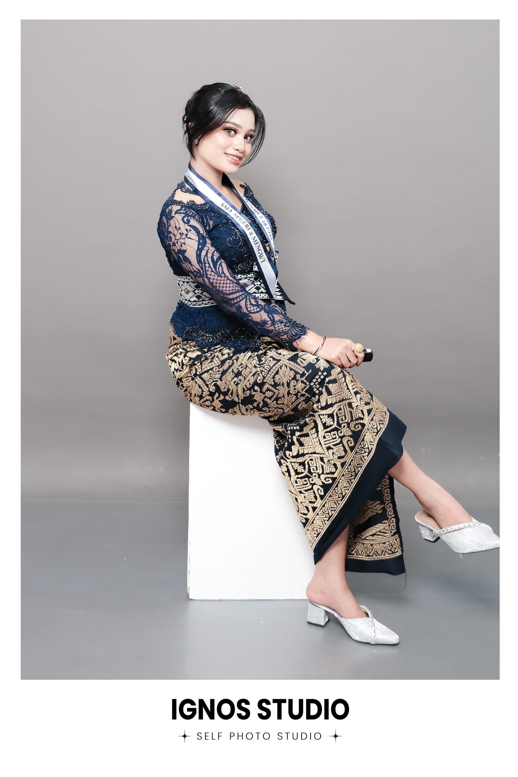Wanita mengenakan kebaya biru modern dan kain batik berpose duduk di Ignos Studio.