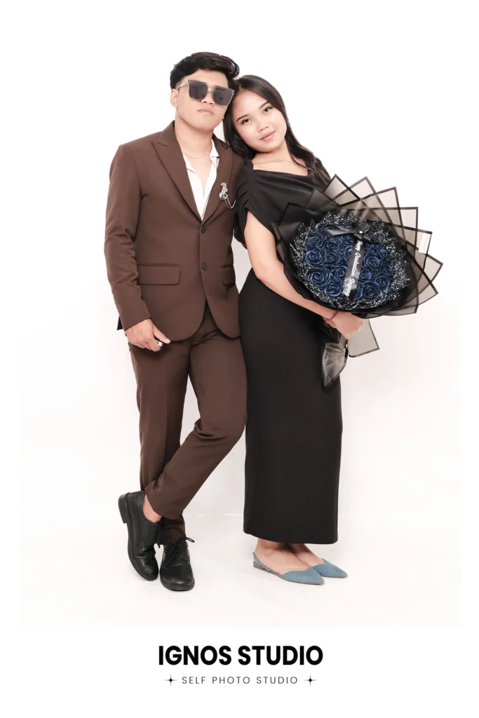 Pasangan berpose formal di Ignos Studio, pria dengan jas cokelat membawa buket bunga besar dan wanita dengan gaun hitam.