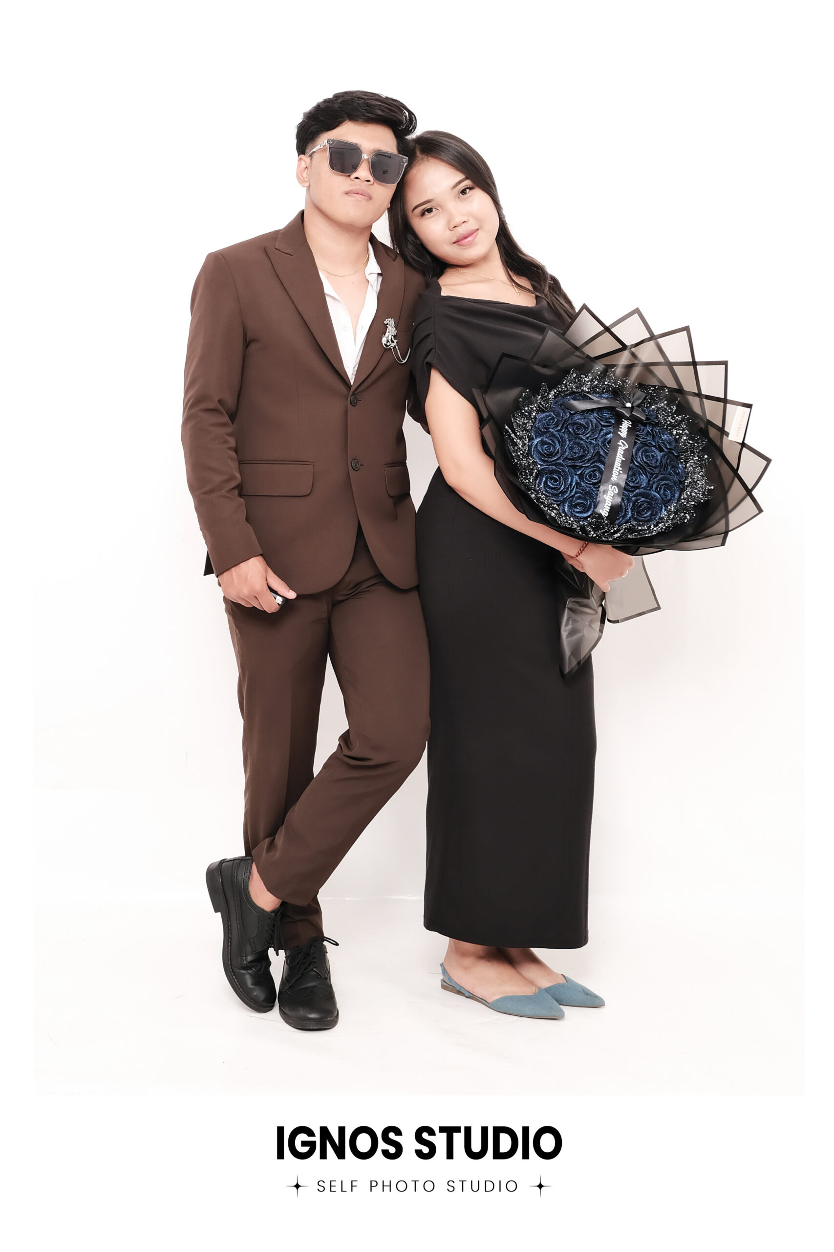 Pasangan berpose formal di Ignos Studio, pria dengan jas cokelat membawa buket bunga besar dan wanita dengan gaun hitam.