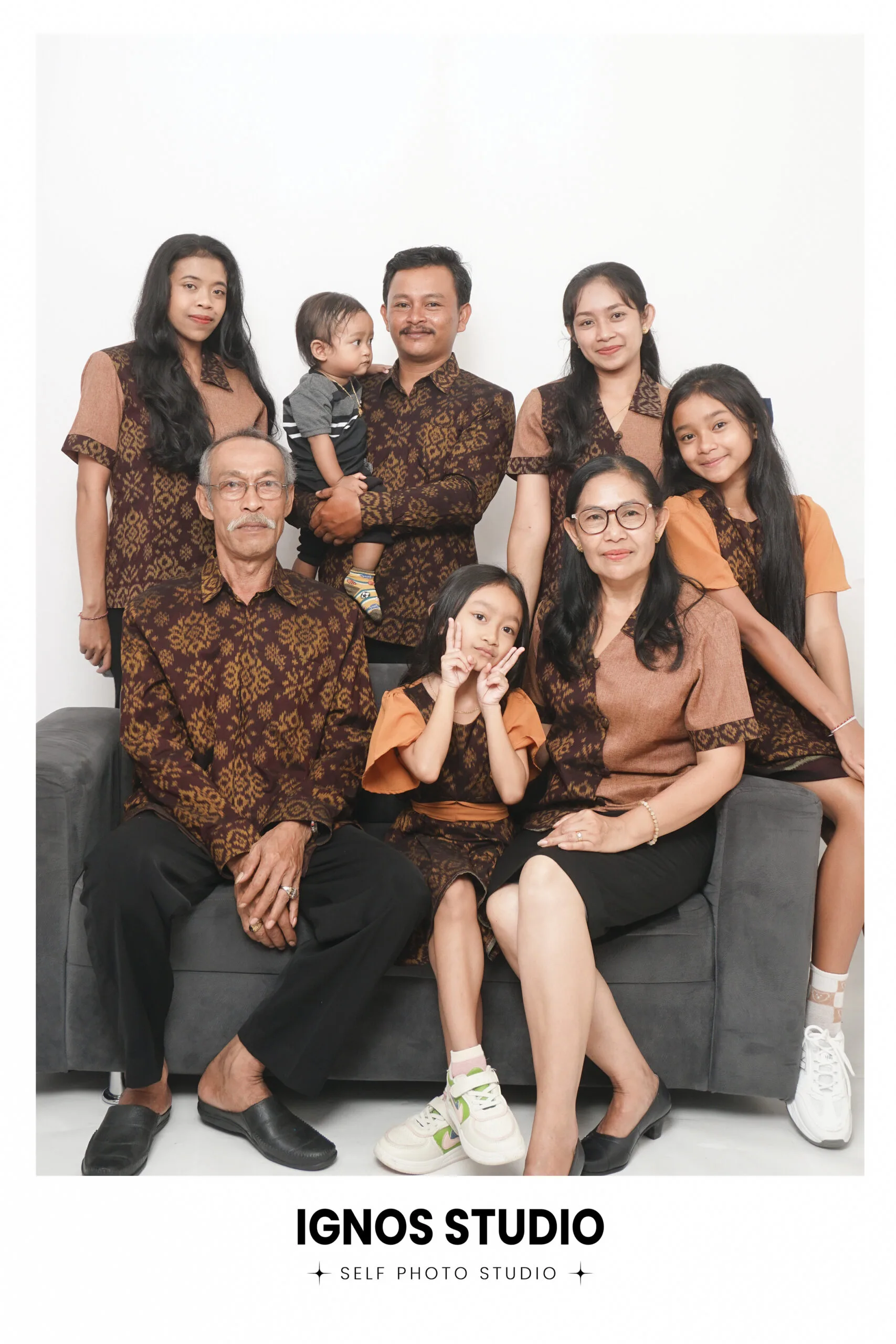 Foto keluarga besar mengenakan seragam batik cokelat seragam berpose di sofa studio Ignos Studio.
